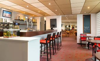 a modern bar with a long wooden counter and several stools , along with various bottles and cups on the shelves at Ibis Longwy Mexy