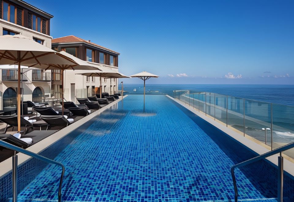 a long , blue pool is surrounded by lounge chairs and umbrellas in front of a building with a view of the ocean at The Setai Tel Aviv, a Member of the Leading Hotels of the World