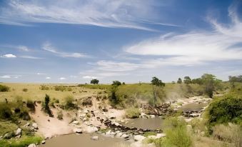 a river flows through a dry , grassy landscape with trees and bushes , under a blue sky dotted with clouds at Mara Intrepids Tented Camp