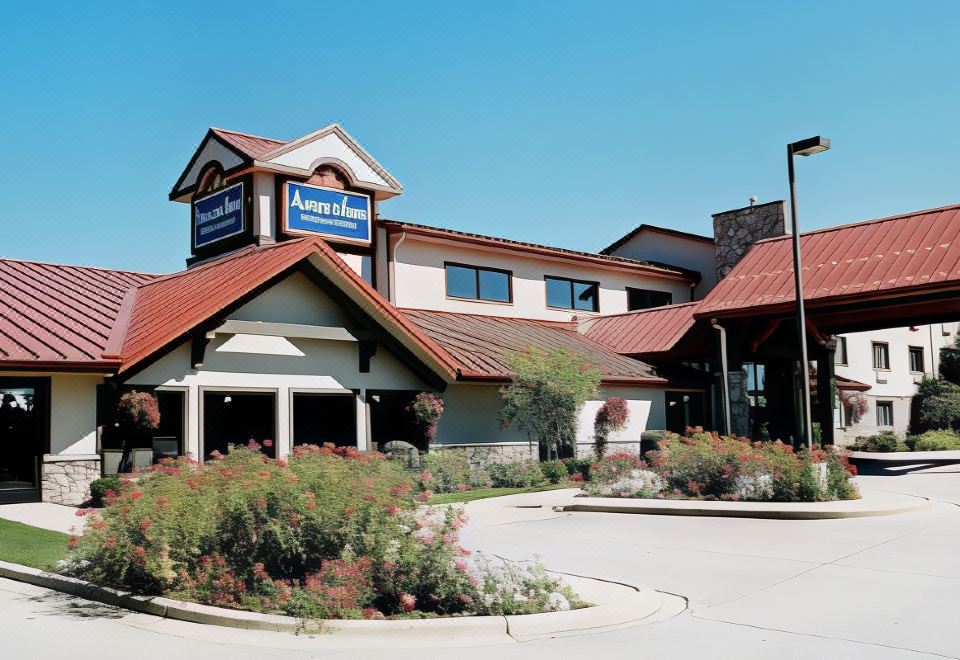 a large hotel building with a red roof and a blue sign on top , surrounded by bushes and flowers at AmericInn by Wyndham Oswego