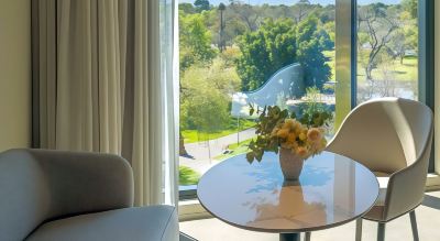 a hotel room with a view of a park , featuring a couch and a dining table with a vase of flowers on it at Oval Hotel at Adelaide Oval, an EVT hotel