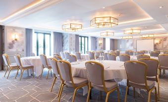 a large conference room with several round tables and chairs , white tablecloths , and multiple pendants hanging from the ceiling at The Billesley Manor Hotel