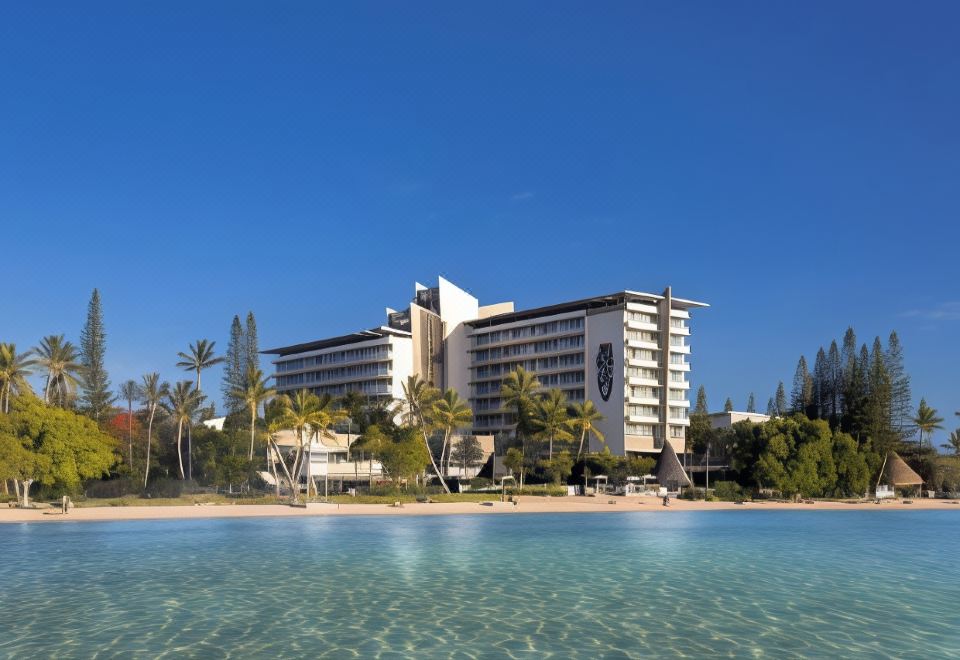 a large white building is situated on the beach with palm trees and a clear blue sky at Chateau Royal Beach Resort & Spa, Noumea