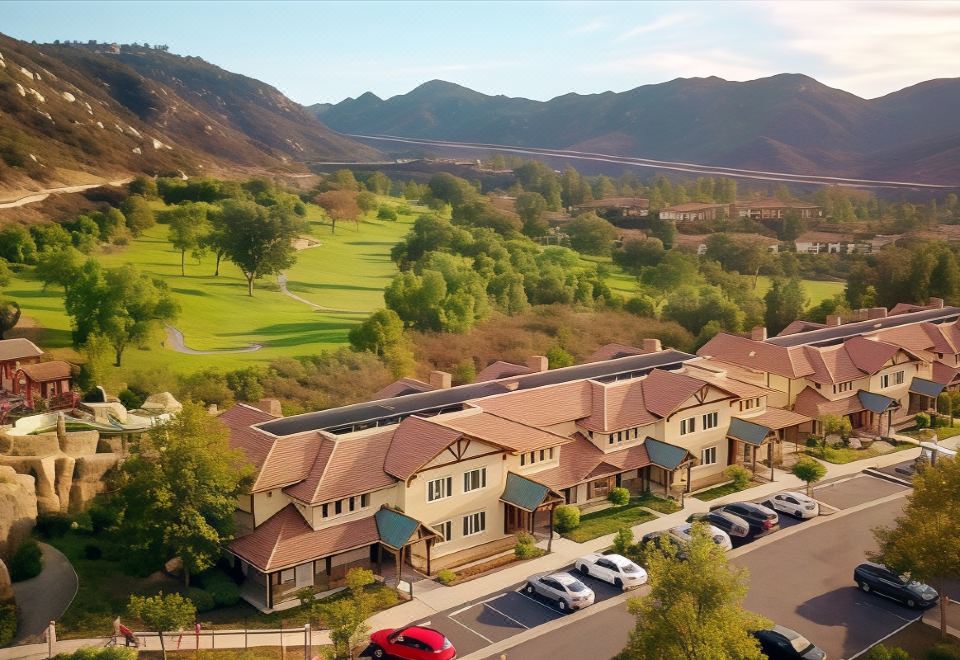 a large , two - story building with a red roof is surrounded by trees and mountains , with cars parked on the street at Hyatt Vacation Club at the Welk, San Diego