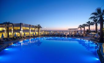 a large outdoor pool surrounded by palm trees , with lounge chairs and umbrellas placed around the pool at Palm Wings Kusadasi Beach Resort&Spa