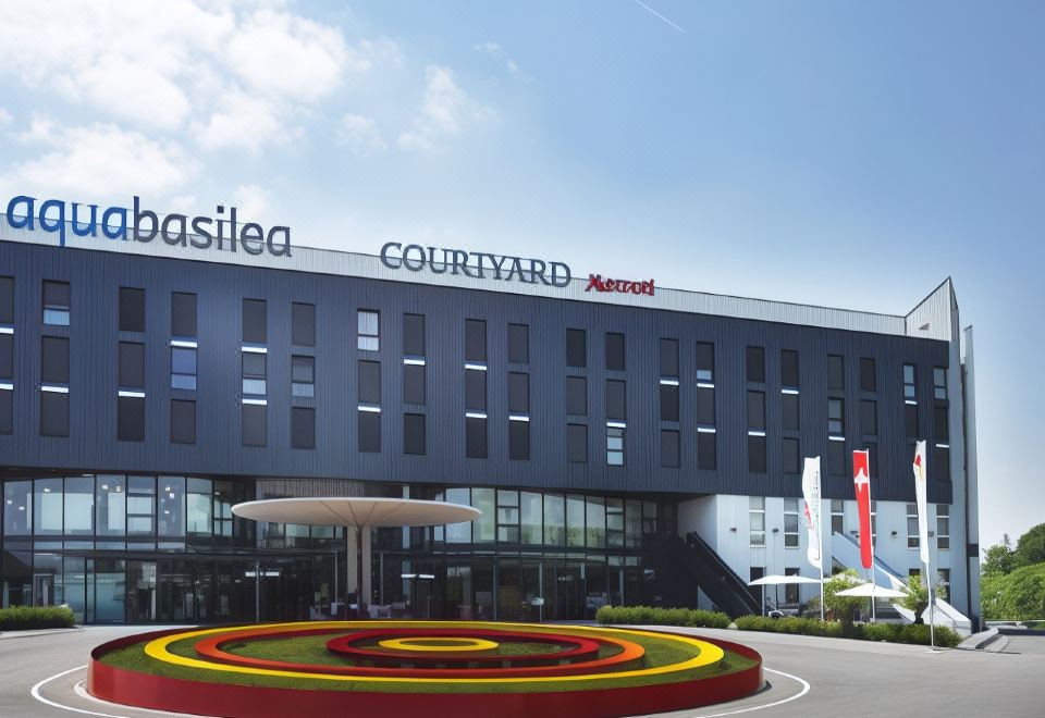 a large hotel with a fountain in front of it , surrounded by grass and trees at Courtyard Basel