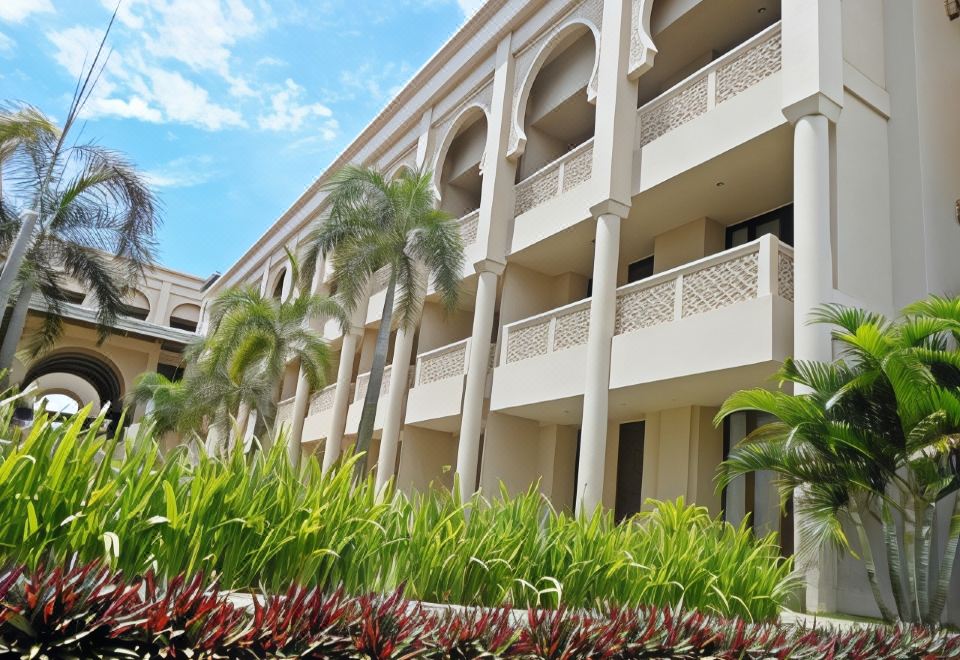 a white building with columns and balconies , surrounded by lush greenery and red flowers , under a clear blue sky at The Pade Hotel