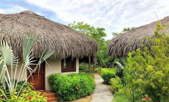 a thatched - roof hut with wooden doors and windows , surrounded by lush greenery and a stone path at Riverside Park Eco Resort