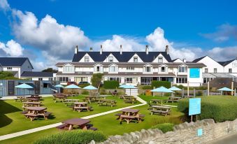 a large , white house surrounded by a grassy field , with several picnic tables and umbrellas in the area at Trearddur Bay Hotel