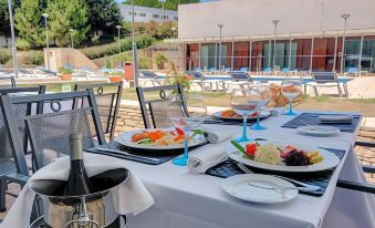 a table is set with plates of food , wine glasses , and a bottle of wine in front of a swimming pool at VIP Executive Santa Iria Hotel