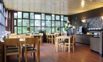 a large , empty dining room with wooden tables and chairs , set against a backdrop of windows and a kitchen area at Bastion Hotel Bussum Hilversum