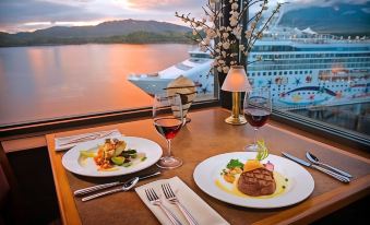 a dining table with two plates of food , wine glasses , and a view of a ship in the background at Crest Hotel