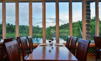 a wooden dining table with chairs and a wine glass is set in front of a large window overlooking a lake at Princess River Kwai Hotel