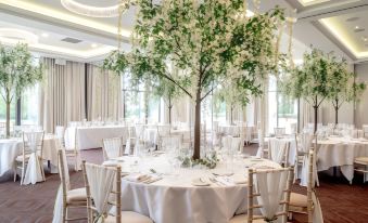 a large , white dining table with multiple chairs and a tree in the center is set for a formal event at Rookery Hall Hotel & Spa
