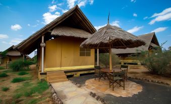 a small wooden cabin with a thatched roof , surrounded by a stone walkway and a table with chairs at AA Lodge Amboseli
