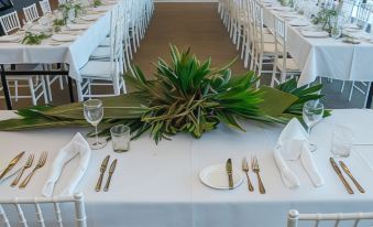 a long dining table is set with white linens , silverware , and a centerpiece of green plants at Caves Coastal Bar & Bungalows