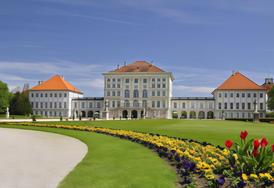 a large white building with a red roof and multiple balconies is surrounded by green grass and flowers at Courtyard Munich Garching