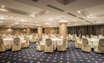 a large , well - lit conference room with white tables and chairs set up for an event , surrounded by blue carpeting and curtains at FlyOn Hotel & Conference Center
