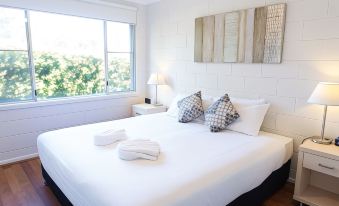 a large bed with white sheets and a lamp on the side is situated in a room with wooden floors and a window at Fingal Bay Holiday Park