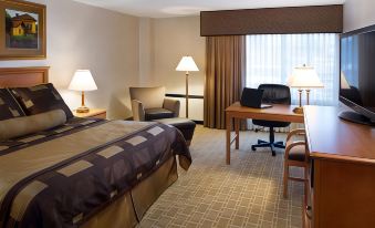 a hotel room with a bed , desk , chairs , and lamps , all set against a backdrop of beige walls and curtains at Bismarck Hotel and Conference Center