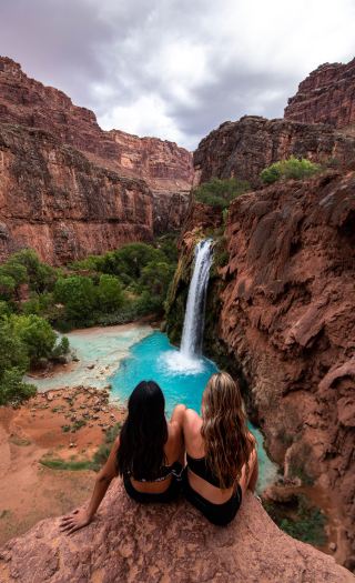Sitting with a view of Havasu Falls, one of many waterfalls on the Havasupai reservation in Arizona, USA. ; Standing in front of Havasu Falls, a bright blue waterfall in the Havasupai reservation in Arizona, USA.