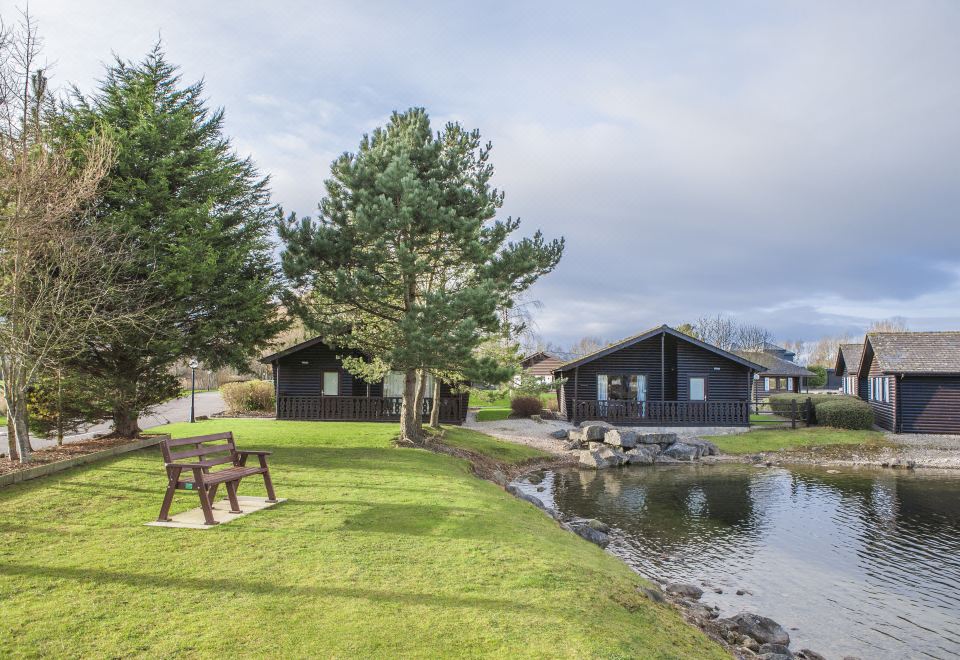 a grassy field with a wooden bench and two small houses situated next to a body of water at Pine Lake Resort