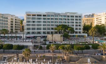 a large , white building with a sculpture on its roof is surrounded by palm trees and people walking around at El Fuerte Marbella