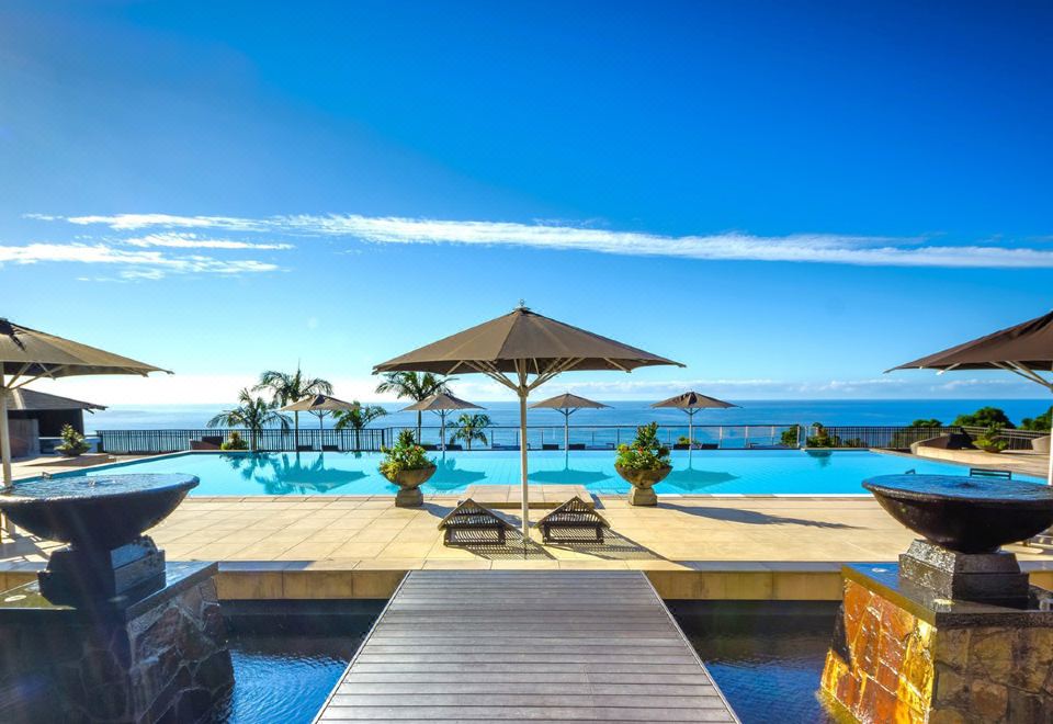 a large swimming pool with a wooden deck and umbrellas , surrounded by palm trees and the ocean at Sankara Hotel & Spa Yakushima