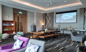 a modern , well - lit conference room with wooden tables and purple chairs , a large screen on the wall , and gray carpeted floor at Grand Fortune Hotel Nakhon Si Thammarat