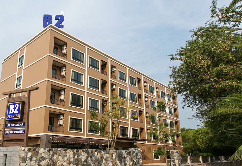 a beige building with a blue sign on the side , surrounded by trees and rocks at B2 Sriracha Premier Hotel