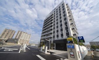 a tall white building with a parking garage on the ground floor is situated next to a road at Just Inn Matsusaka Station