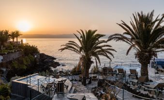 a beautiful sunset over the ocean , with palm trees and rocks in the foreground , and chairs and tables set up on a patio at Catalonia Punta del Rey