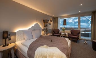 a large bed with a brown blanket and white pillows is in a room with two chairs and a window at Sunstar Hotel & Spa Grindelwald