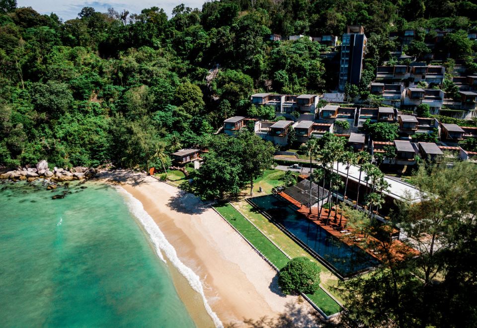 aerial view of a sandy beach near a city , with trees and buildings in the background at The Naka Phuket, a member of Design Hotels