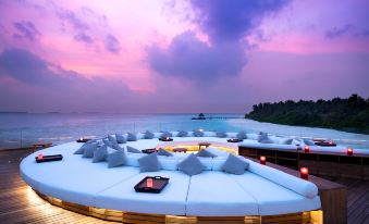 a large outdoor seating area with multiple couches and chairs , overlooking the ocean at sunset at Anantara Kihavah Maldives Villas
