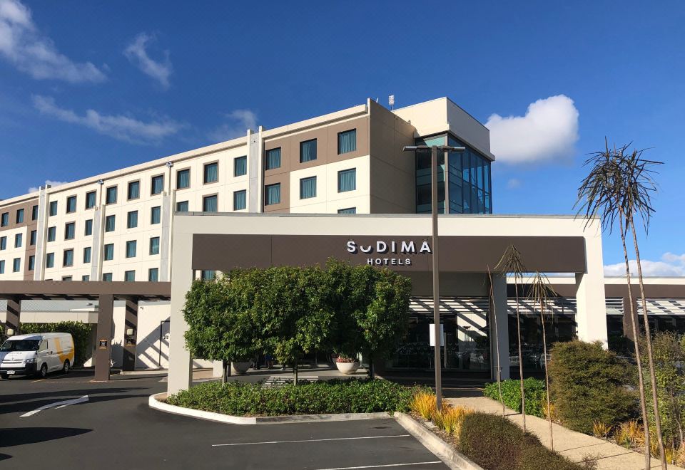 a large hotel with a parking lot in front of it , surrounded by trees and grass at Sudima Auckland Airport