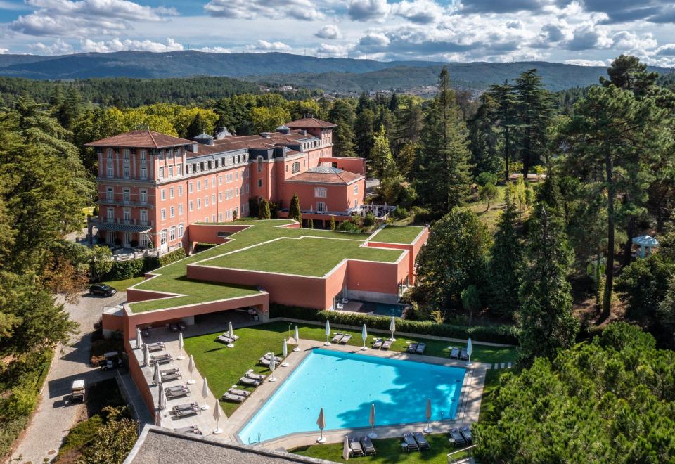 a large hotel surrounded by green grass and trees , with a swimming pool in the foreground at Vidago Palace