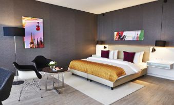 a large bed with a gold blanket is in the middle of a room with a black chair , table , and paintings at Steigenberger Hotel Köln