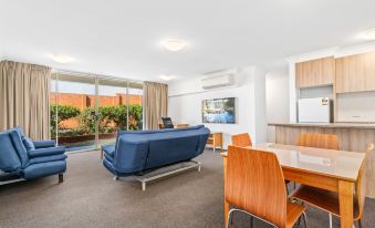 a modern living room with a blue couch , wooden table , and chairs , along with large windows and a kitchen area at APX Parramatta