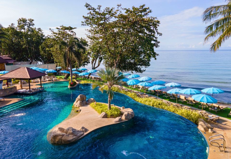 a tropical resort with a pool , palm trees , and blue umbrellas near the ocean , all set against a backdrop of clear skies at Kacha Resort & Spa, Koh Chang