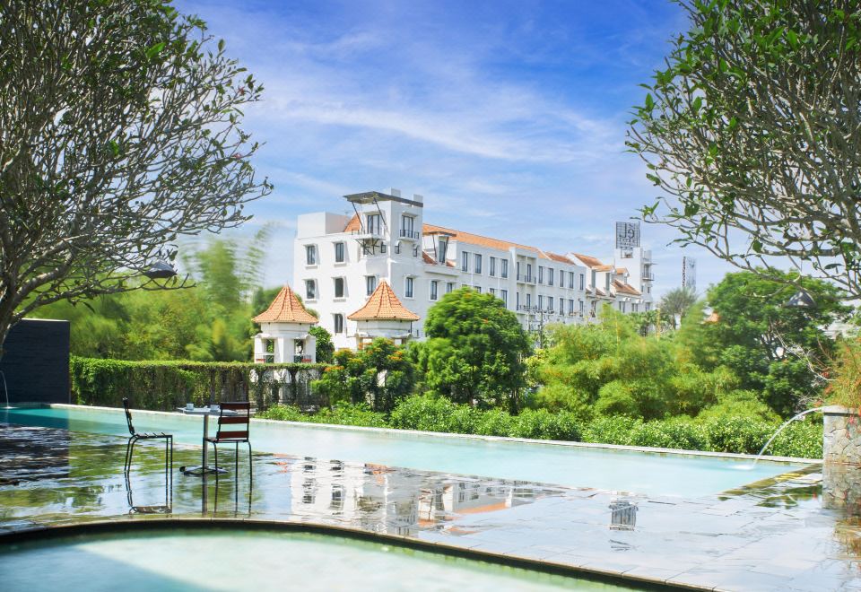 a large white building with a brown roof is surrounded by trees and a reflecting pool at El Hotel Malang