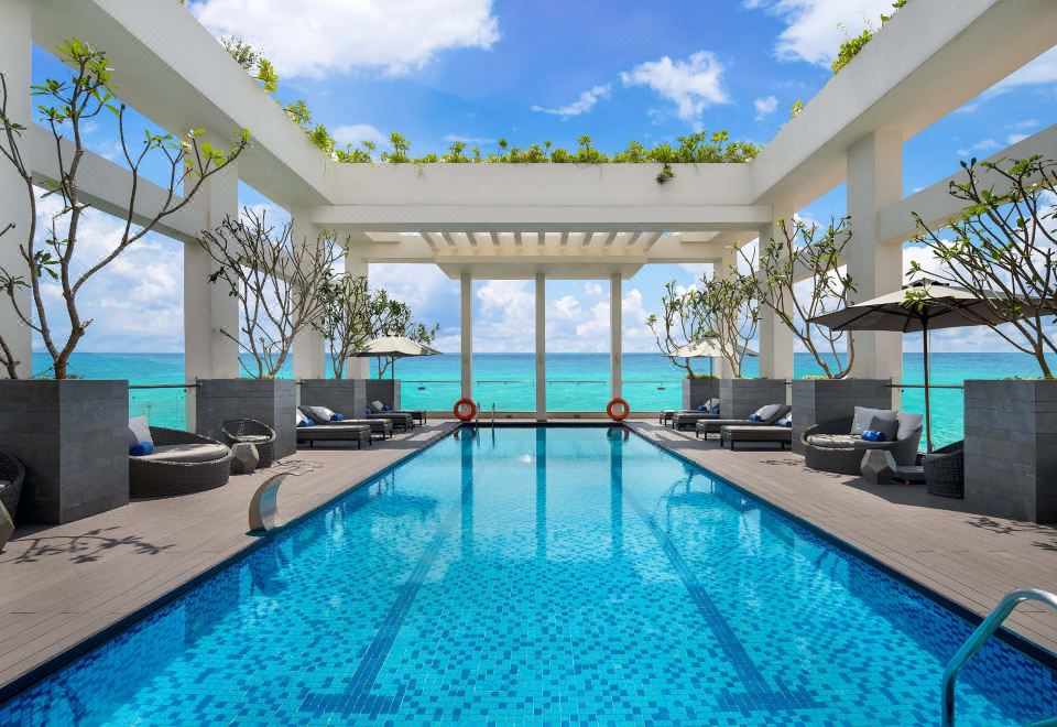 an outdoor pool surrounded by lounge chairs and umbrellas , with a view of the ocean in the background at Paris Deli Danang Beach Hotel