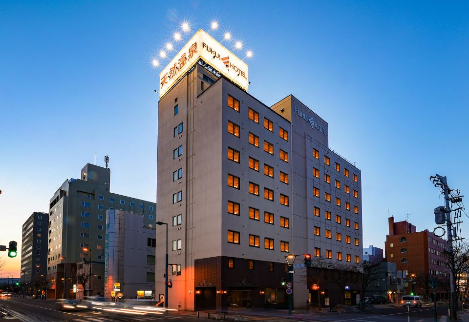 a modern building with a large sign above the entrance , illuminated at night , and surrounded by other buildings in a city setting at Fukui Hotel