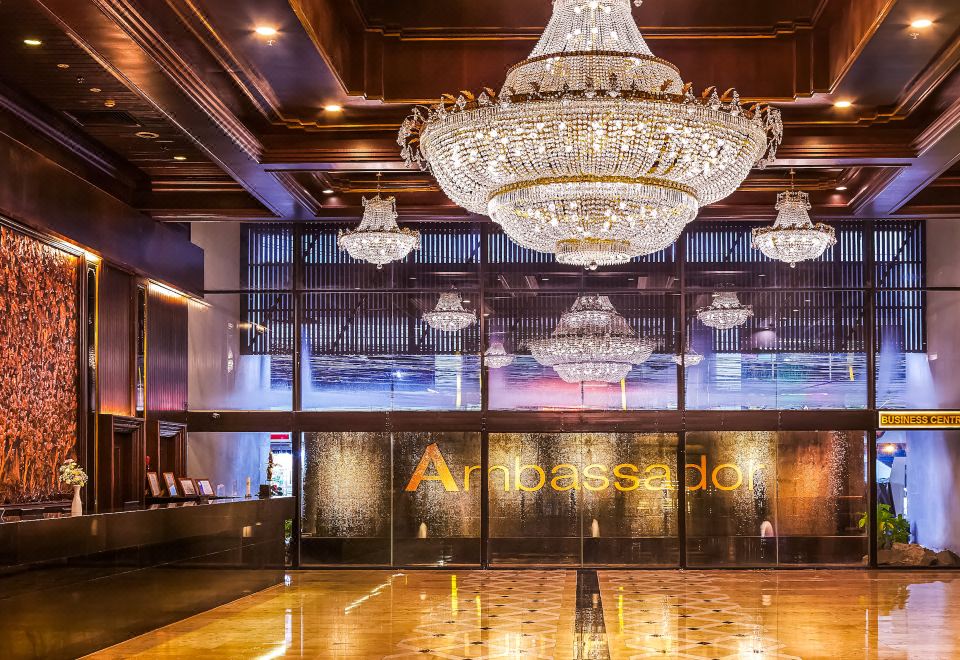 A large chandelier hangs elegantly from the ceiling of a lobby adorned with multiple windows and wooden accents at Ambassador Hotel Bangkok