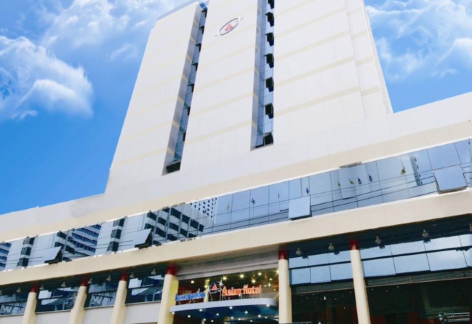 a tall white building with a red circle logo is surrounded by trees and potted plants at Asian Hotel