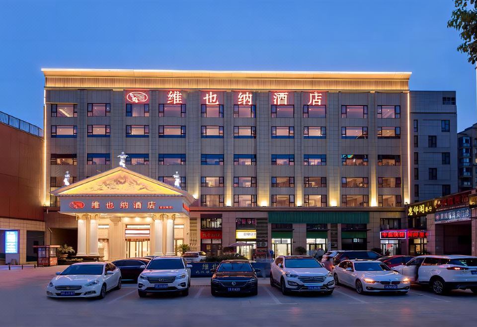 a large hotel with a red and white facade is lit up at night , with several cars parked in front of it at Vienna Hotel