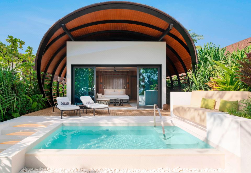 a small pool is situated next to a wooden building with an arched roof , surrounded by lush greenery at The Westin Maldives Miriandhoo Resort