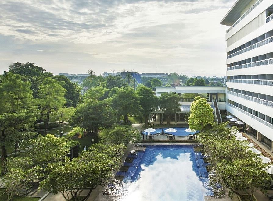 a large swimming pool is surrounded by trees and buildings , with the sky above appearing hazy at Royal Ambarrukmo Yogyakarta