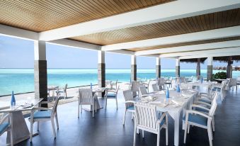 a large , modern restaurant with white tables and chairs arranged on a patio overlooking the ocean at Anantara Dhigu Maldives