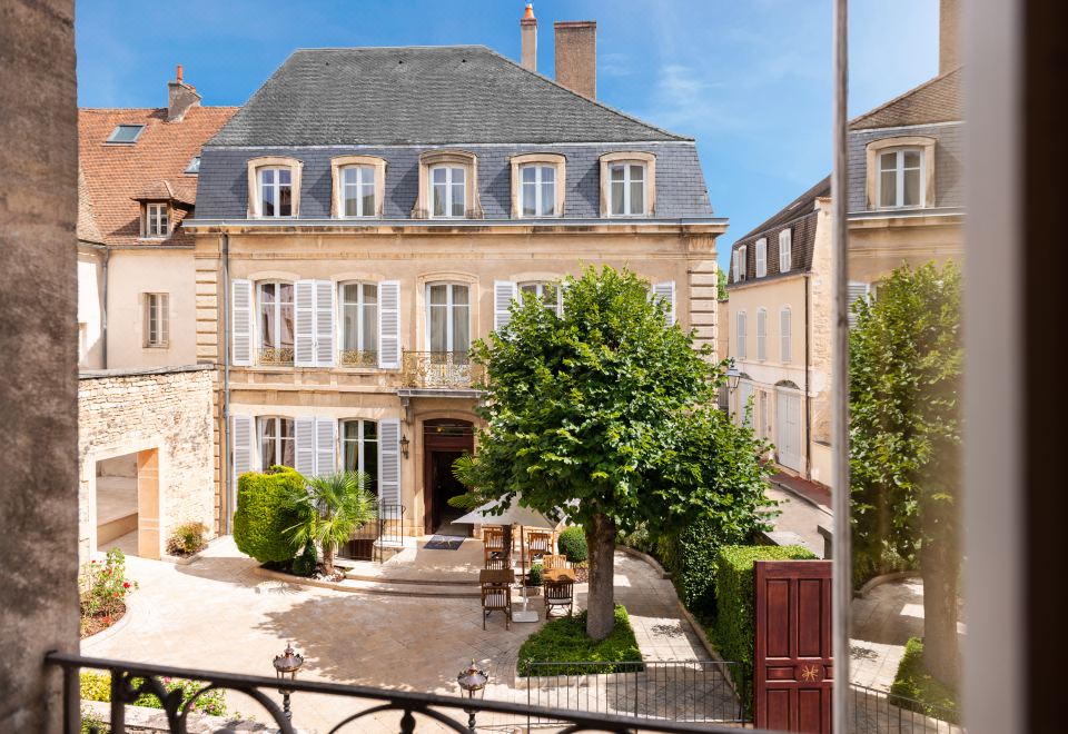 a large , two - story building with a stone facade and white shutters is surrounded by trees and a patio area at L'Hotel de Beaune